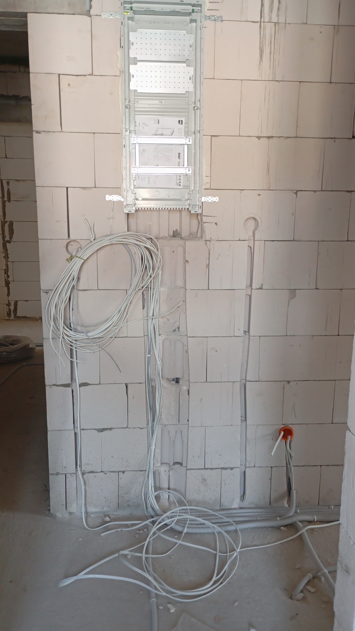Rozdzielnica elektryczna zamontowana na ścianie z betonu komórkowego. Widoczne kable w bruzdach i na podłodze. Etap prac instalacyjnych w surowym stanie.