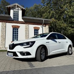 Ds-cars - Biały Renault Megane sedan na tle budynku z ceglanymi detalami i zieleni. Auto zaparkowane na kostce brukowej, widoczne tablice rejestracyjne. Słoneczny dzień.
