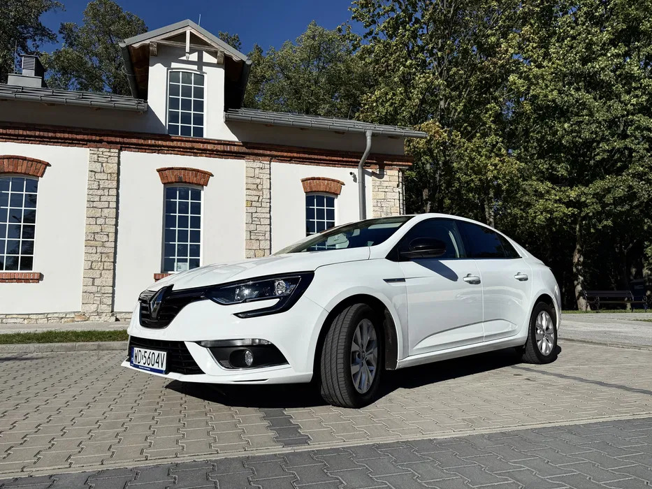 Biały Renault Megane sedan na tle budynku z ceglanymi detalami i zieleni. Auto zaparkowane na kostce brukowej, widoczne tablice rejestracyjne. Słoneczny dzień.
