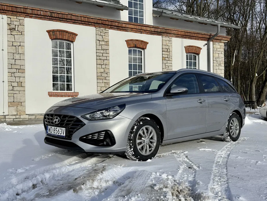 Srebrny Hyundai i30 kombi na śniegu przed budynkiem z ceglanymi detalami. Zimowe opony. Wynajem aut na zimowe warunki.