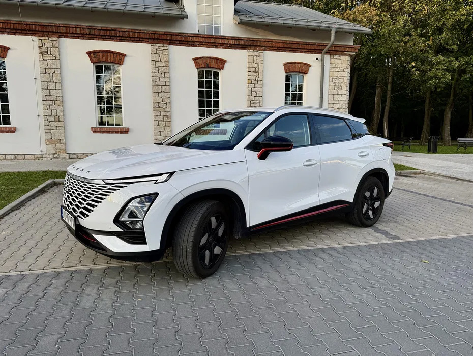 Biały crossover z czarnymi felgami i czerwonym akcentem, zaparkowany na kostce brukowej przed budynkiem z ceglanymi detalami. Nowoczesny design, widok z boku.