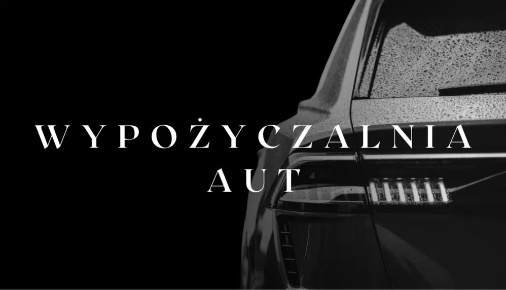 Czarno-białe zdjęcie tyłu samochodu z napisem 'Wypożyczalnia aut'. Widoczne krople deszczu na karoserii i charakterystyczne światła tylne.