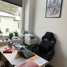 MoniCo Monika Kozłowska - Biuro z laptopem, dokumentami i krzesłem gamingowym. Na ścianie wisi grafika z motywem miejskim. Widok z okna na ośnieżone budynki i drzewa.