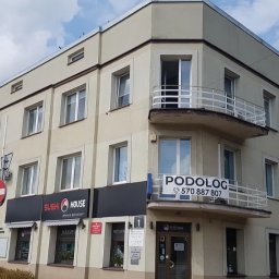 MoniCo Monika Kozłowska - Trzypiętrowy budynek z balkonami, szyldem 'PODOLOG' i 'SUSHI HOUSE'. Widoczne znaki drogowe i zieleń. Ujęcie z poziomu ulicy w słoneczny dzień.
