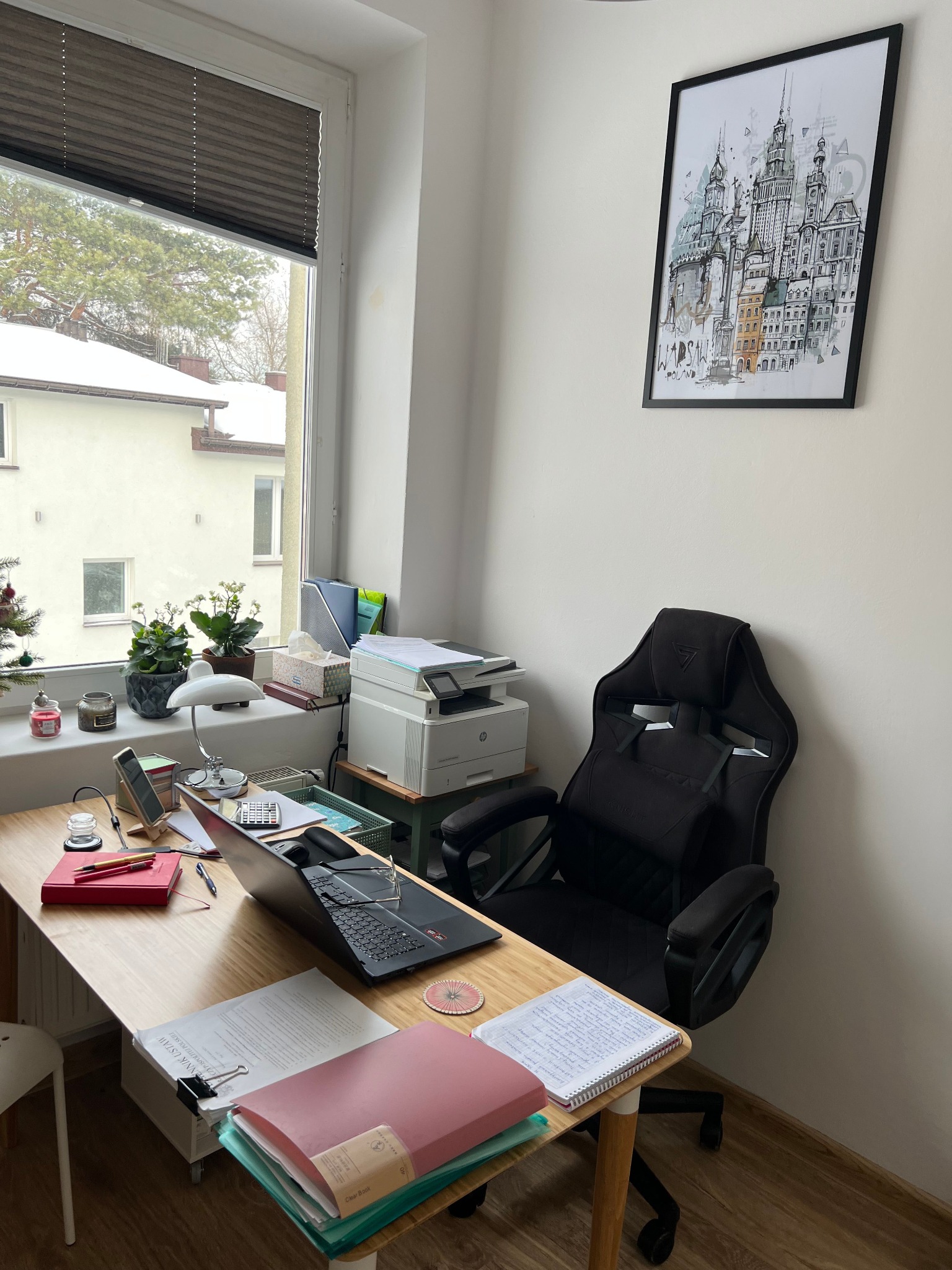 Biuro z laptopem, dokumentami i krzesłem gamingowym. Na ścianie wisi grafika z motywem miejskim. Widok z okna na ośnieżone budynki i drzewa.
