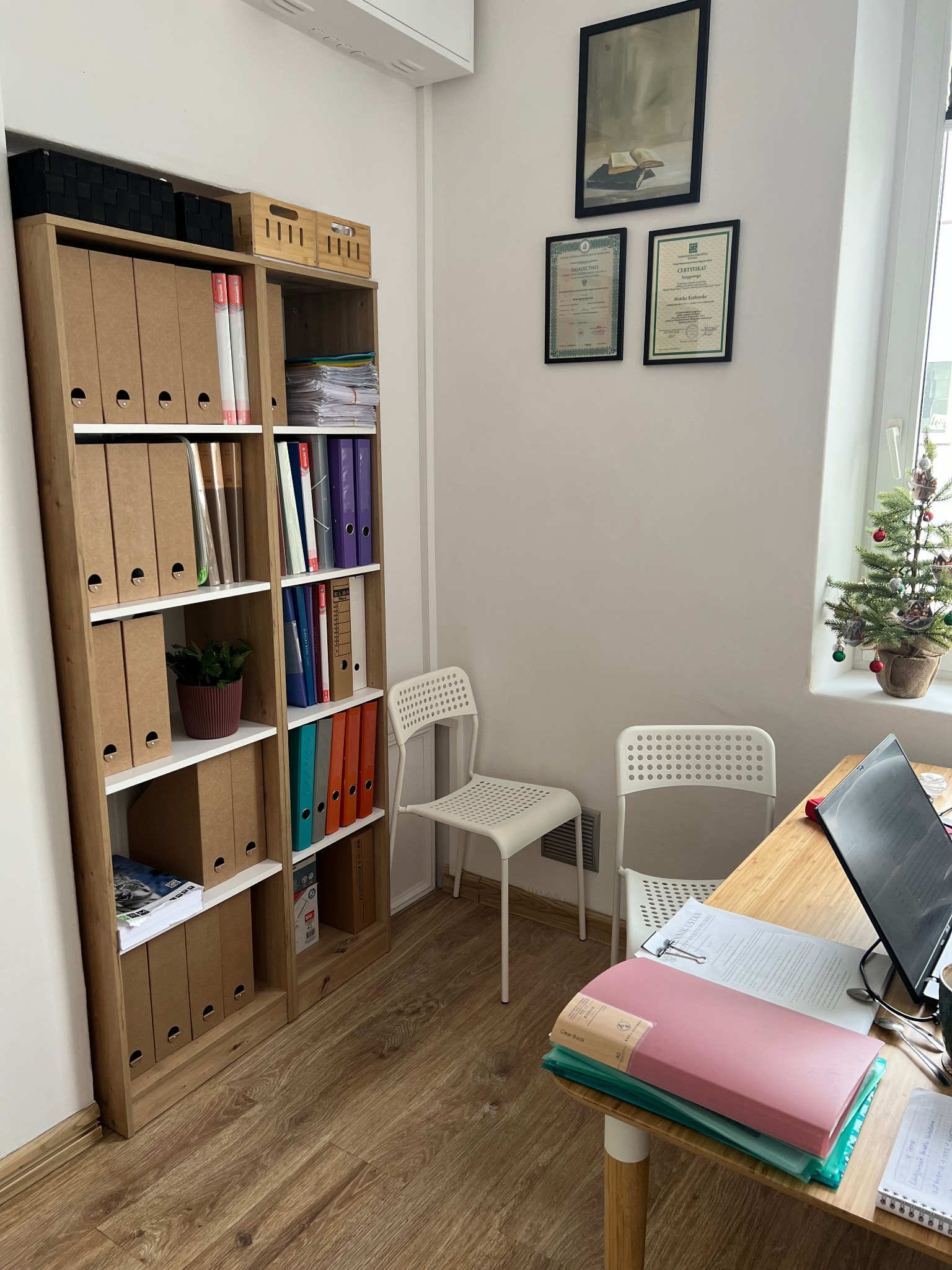 Biuro w Warszawie: regał z segregatorami, certyfikaty na ścianie, biurko z dokumentami i komputerem. W tle okno z małą choinką.