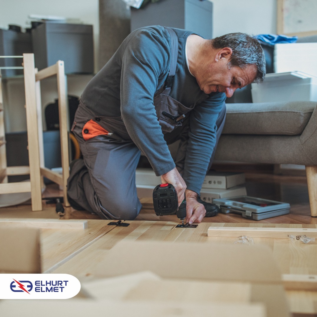 Montażysta skręca drewniane elementy mebli w pomieszczeniu z kartonami. Używa wkrętarki akumulatorowej. Widoczne fragmenty sofy i podłogi.