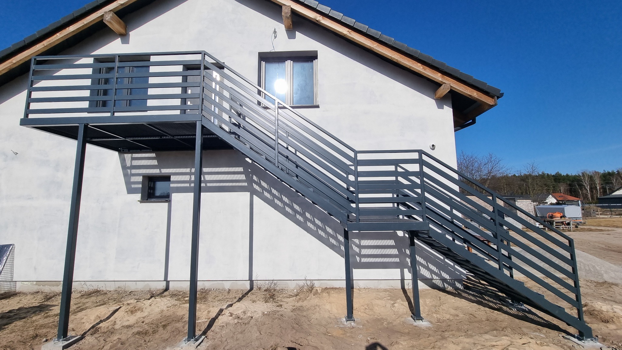 Zewnętrzna, metalowa konstrukcja schodów z balustradą prowadząca na balkon domu jednorodzinnego. Nowoczesny design, szara kolorystyka, widok z dołu.