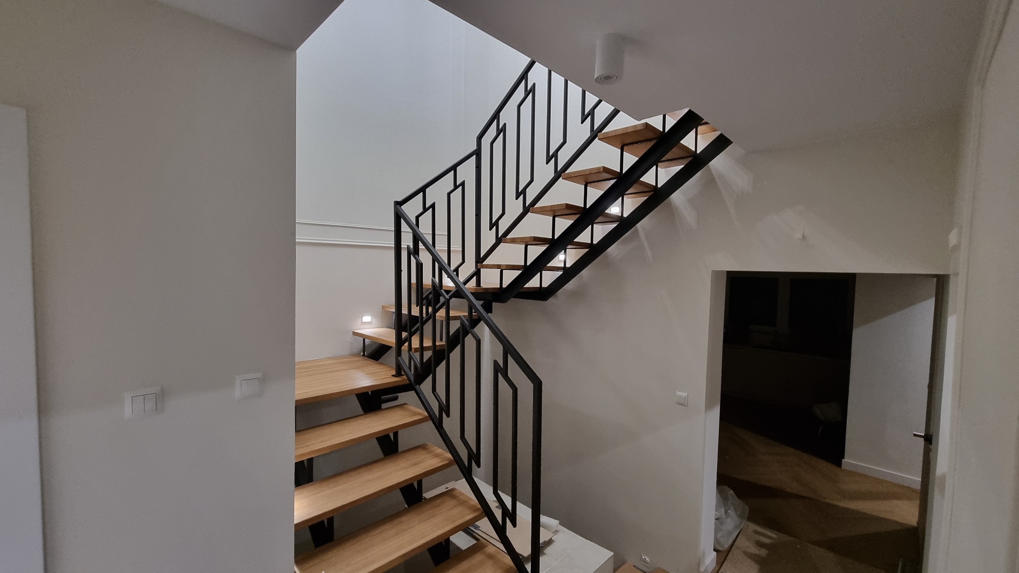 Nowoczesne schody z drewnianymi stopniami i czarną, geometryczną balustradą metalową, oświetlone punktowo, w jasnym wnętrzu domu. Minimalistyczny design.