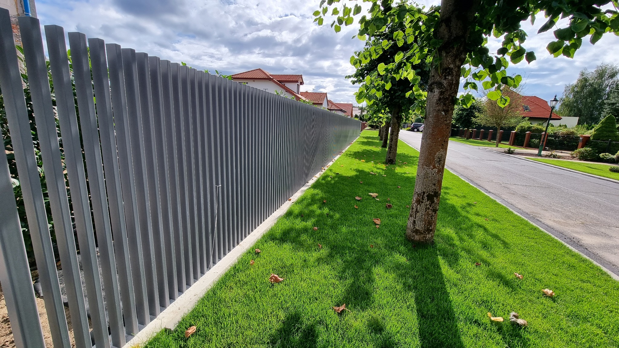 Nowoczesne, pionowe ogrodzenie metalowe w kolorze szarym, oddzielające posesję od ulicy. Widoczne drzewa i domy w tle, trawnik z opadłymi liśćmi. Słoneczny dzień.