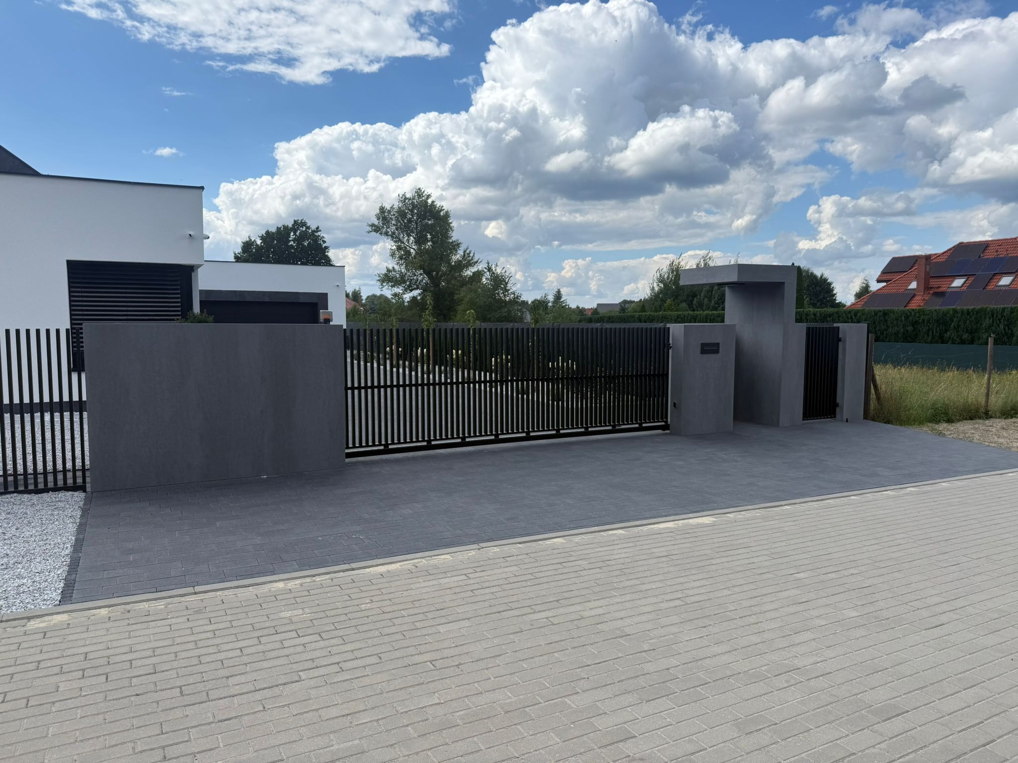 Nowoczesna, czarna brama wjazdowa z pionowymi szczeblami i szarymi, betonowymi słupkami w Solcu Kujawskim. Minimalistyczny design, błękitne niebo w tle.