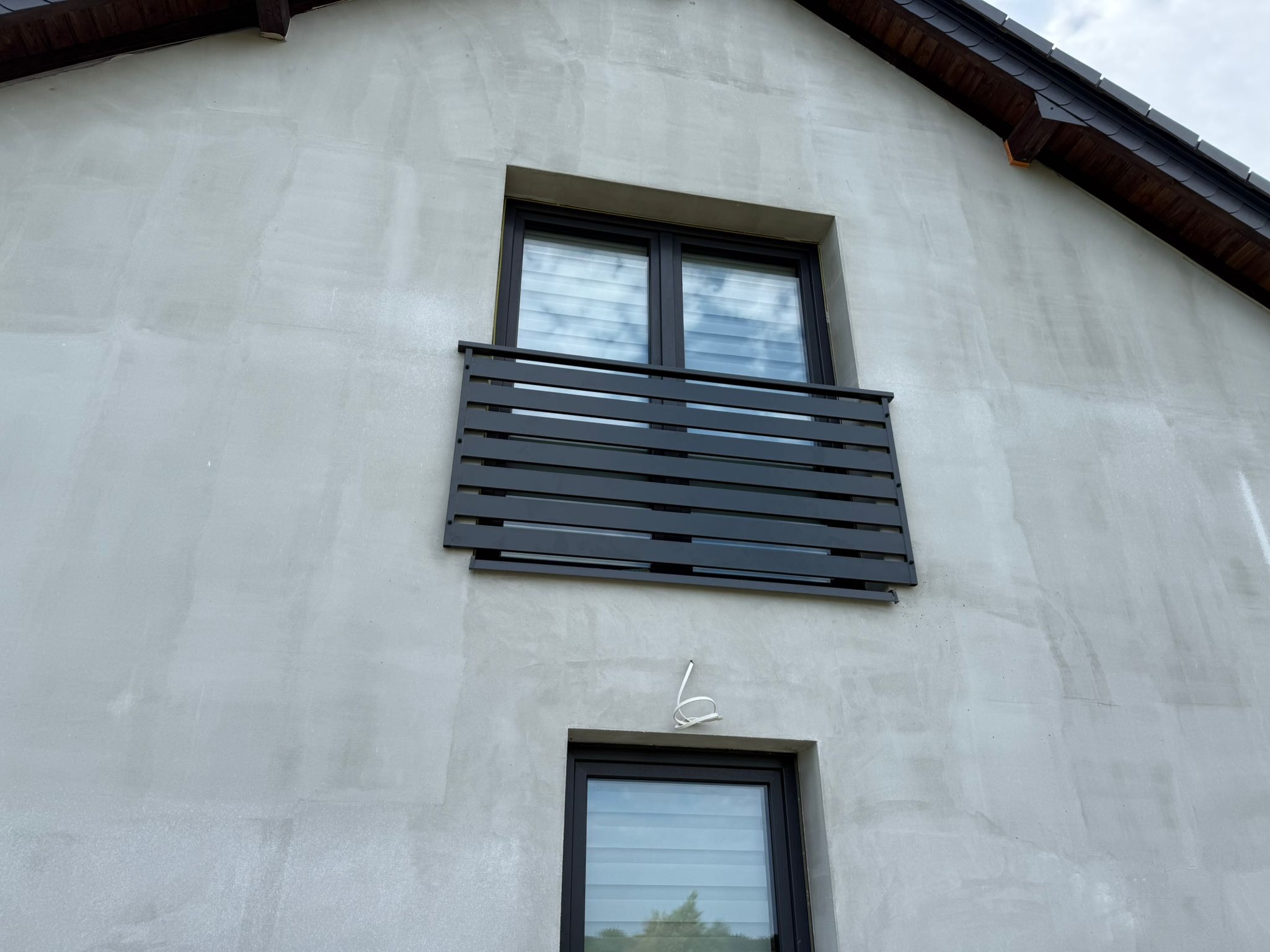 Szara, pozioma balustrada balkonowa z metalowych profili, zamontowana na elewacji domu z dwoma oknami. Widok z dołu, na tle pochmurnego nieba.