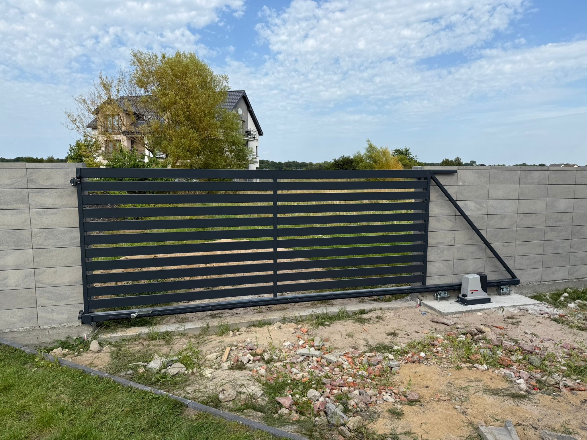 Nowoczesna, przesuwna brama wjazdowa z automatyką, z poziomymi panelami, zamontowana przy ogrodzeniu z szarej kostki. W tle dom i drzewa pod błękitnym niebem.