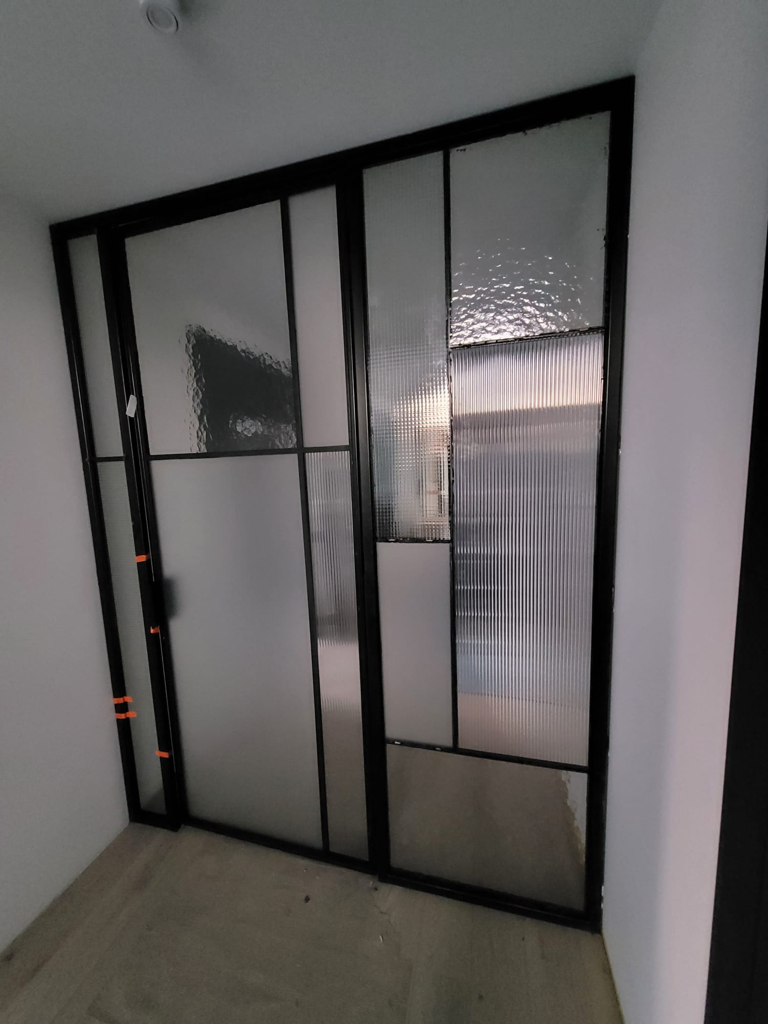 Nowoczesne drzwi przesuwne z czarną ramą i szkłem o różnej fakturze, wkomponowane w jasne wnętrze. Minimalistyczny design z geometrycznymi podziałami.