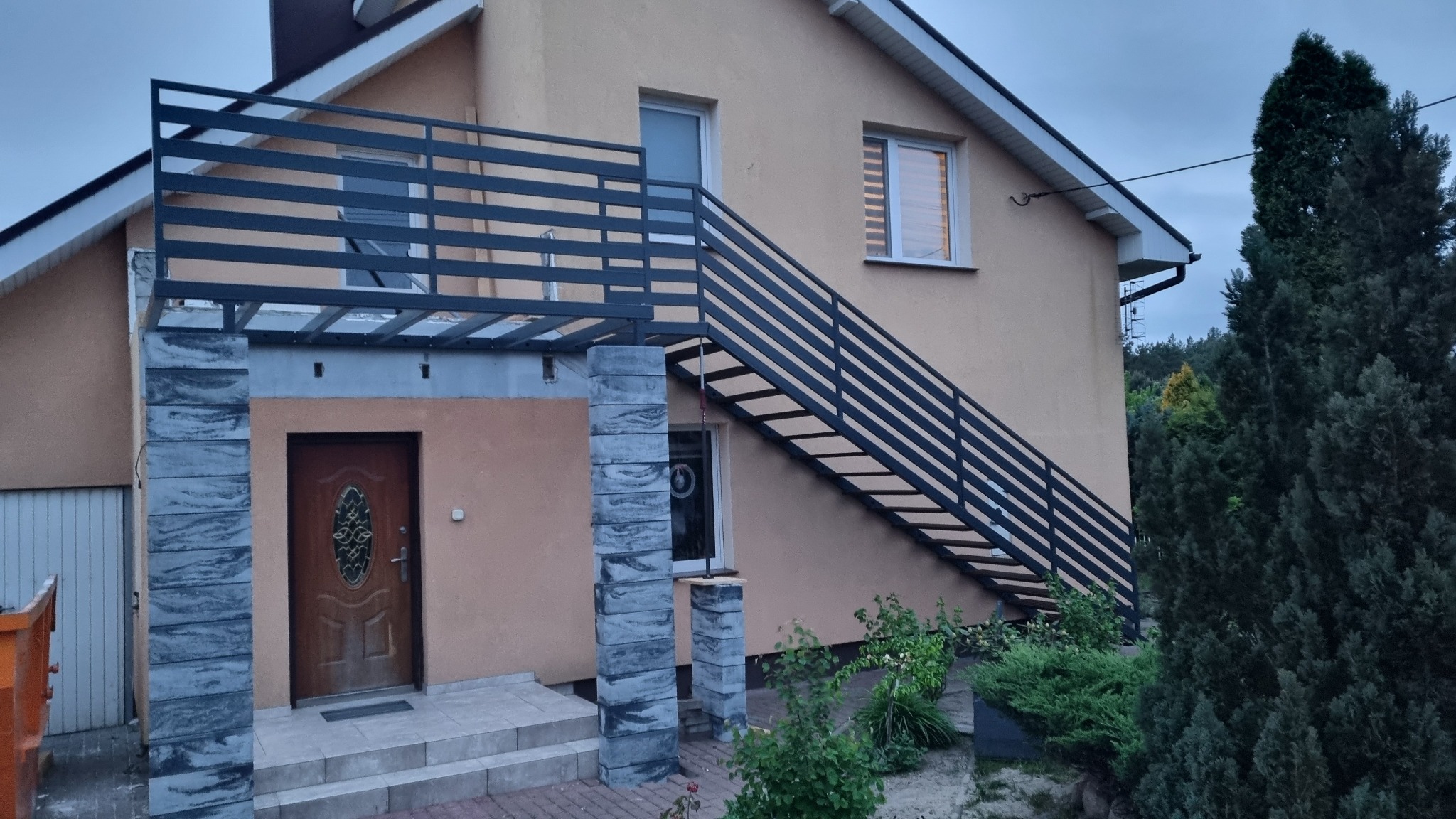 Zewnętrzne schody z czarną balustradą prowadzące na balkon domu z beżową elewacją. Widoczne kolumny z szarej kostki i drzwi wejściowe. Ujęcie z dołu, pochmurne niebo.