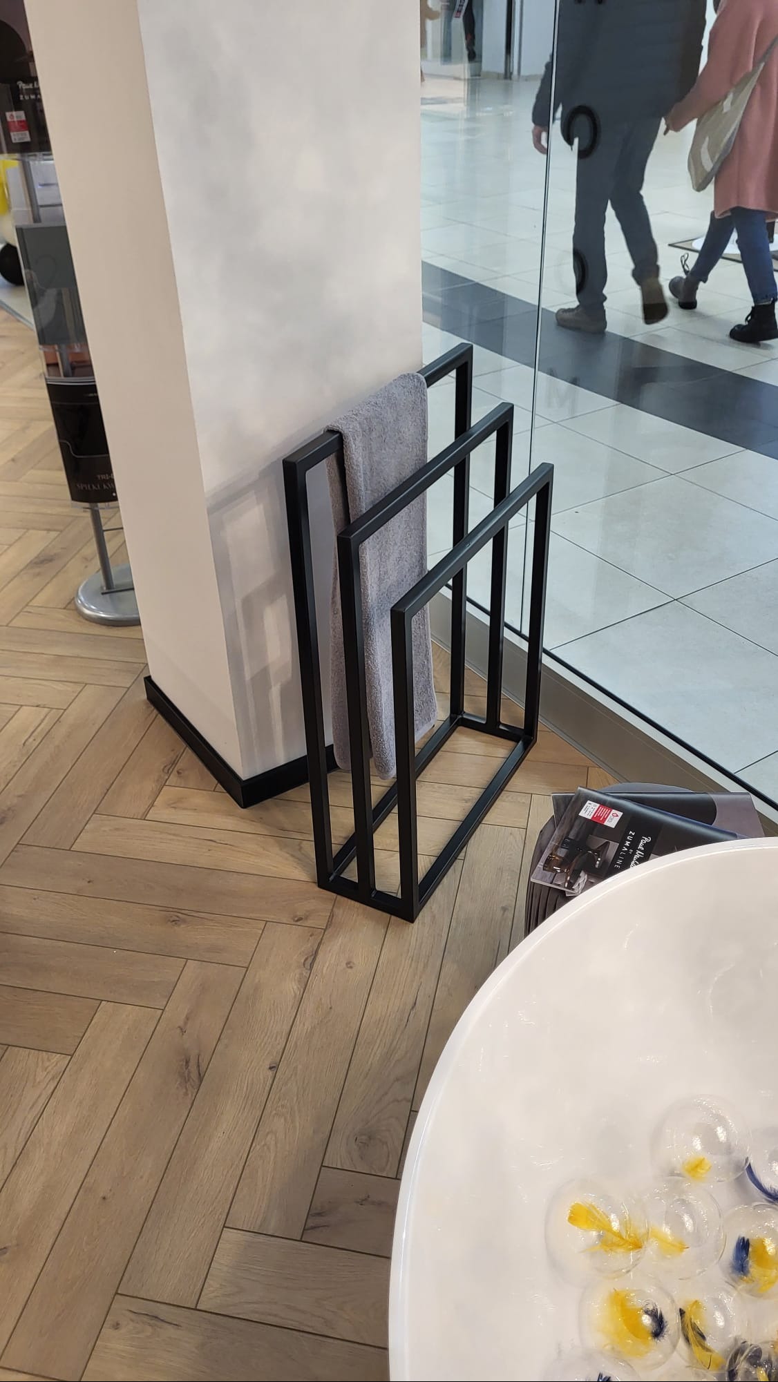 Minimalistyczny, czarny, metalowy stojak na ręczniki z szarą tkaniną, ustawiony obok białej wanny z dekoracyjnymi kulami w jasnym wnętrzu sklepu.