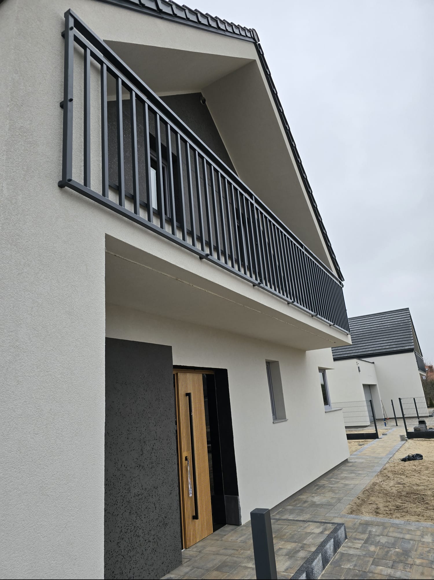 Nowoczesny dom z balkonem zabezpieczonym czarną, metalową balustradą. Minimalistyczny design, jasna elewacja i geometryczne kształty. Widok z dołu, pochmurne niebo.