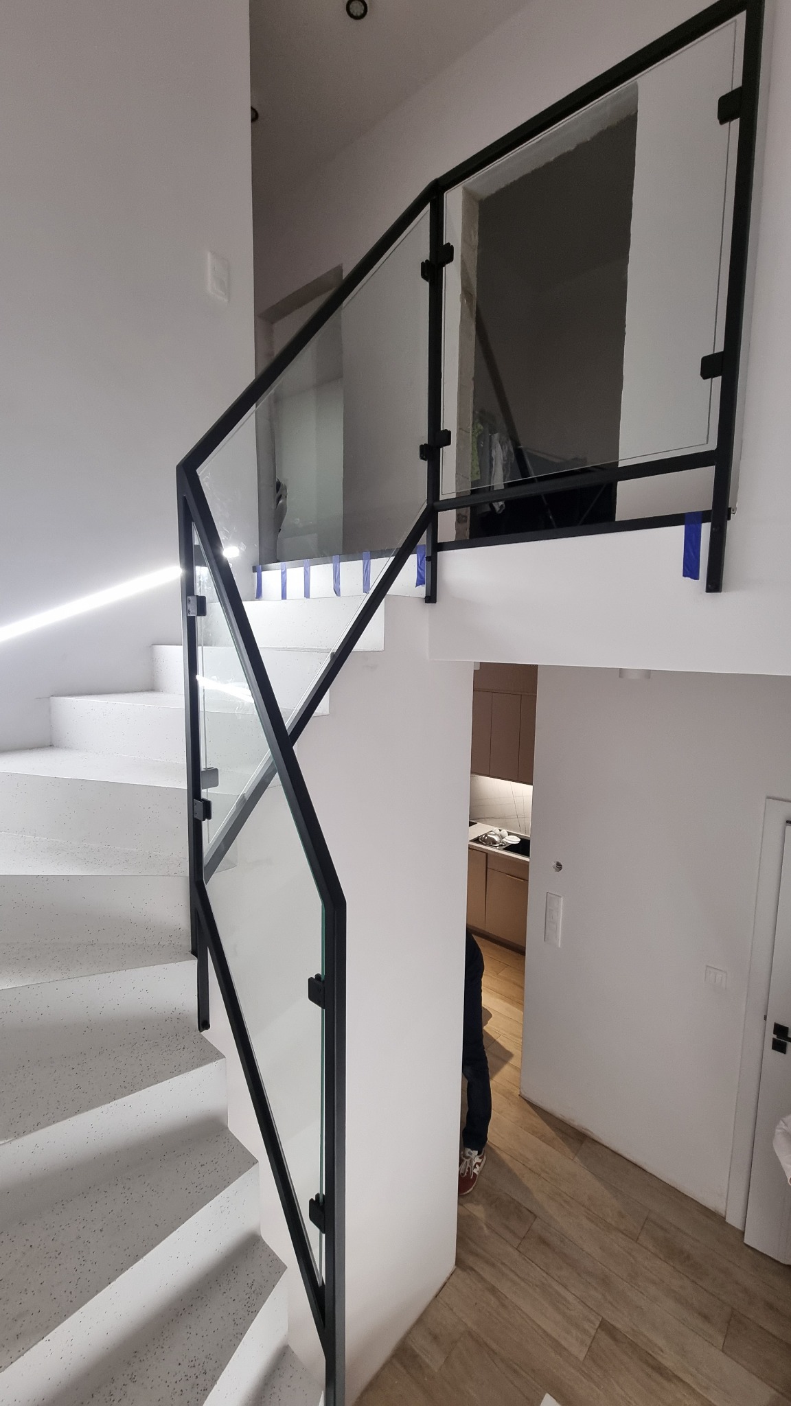 Nowoczesna balustrada szklana z czarnymi metalowymi elementami, zamontowana przy białych schodach w jasnym wnętrzu domu. Widok na otwartą przestrzeń.