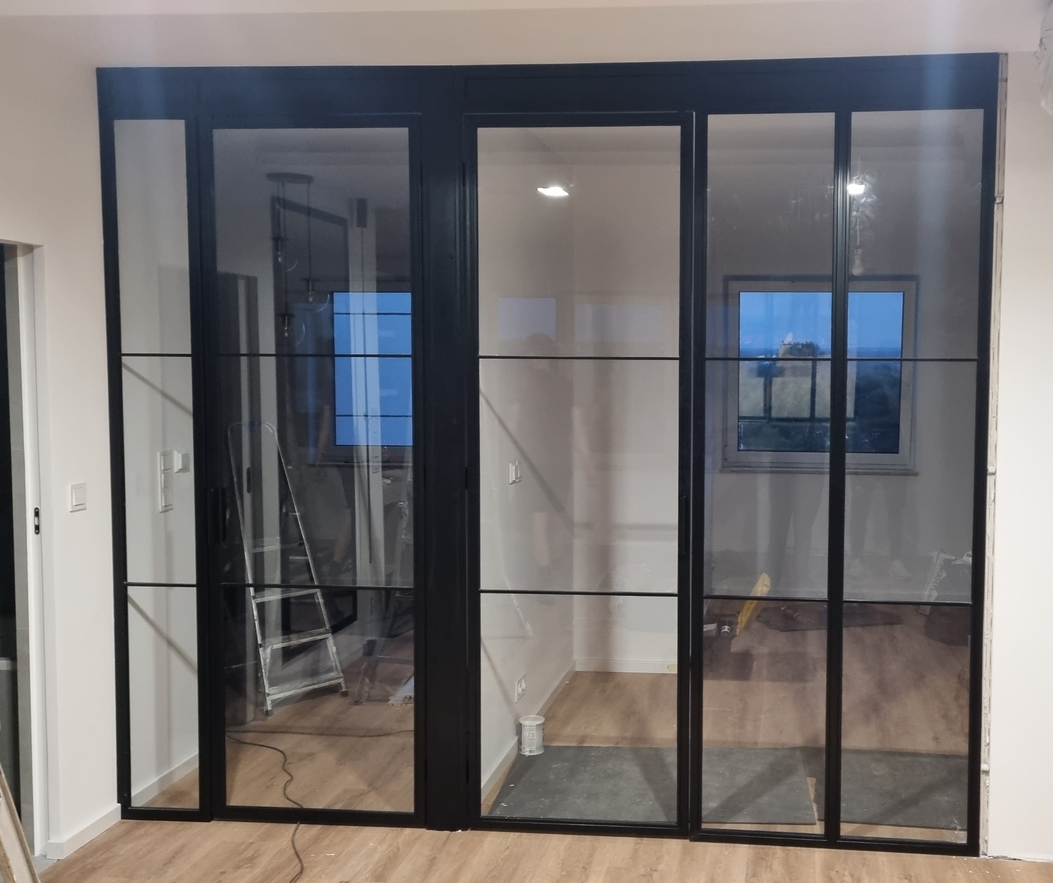 Nowoczesne, przeszklone drzwi przesuwne z czarnymi szprosami, oddzielające pomieszczenia. Widoczny fragment drabiny i narzędzia, sugerujące prace wykończeniowe.