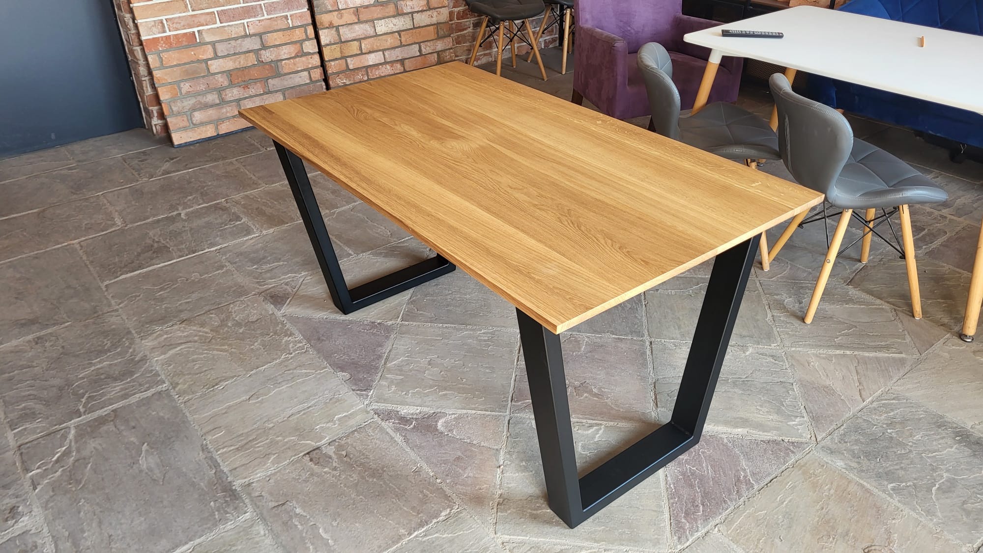 Stół z blatem z jasnego drewna i czarnymi, trapezowymi nogami. Widoczne krzesła i fragment ceglanej ściany w tle. Ujęcie z perspektywy.