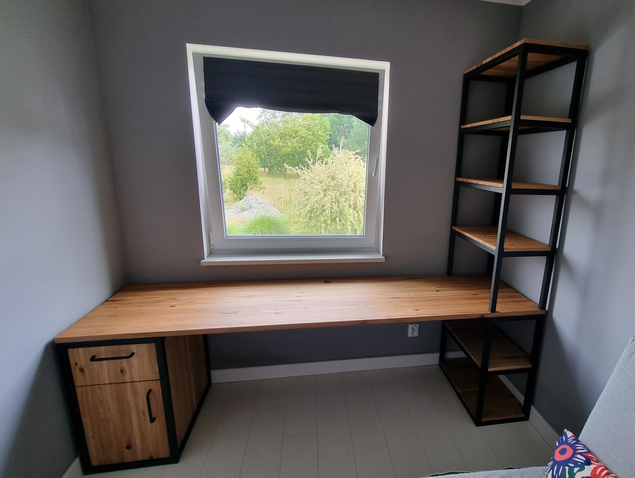 Drewniany blat biurka z metalowym stelażem, szafką i regałem, ustawiony pod oknem z widokiem na zieleń. Nowoczesny design, szare ściany, jasna podłoga.