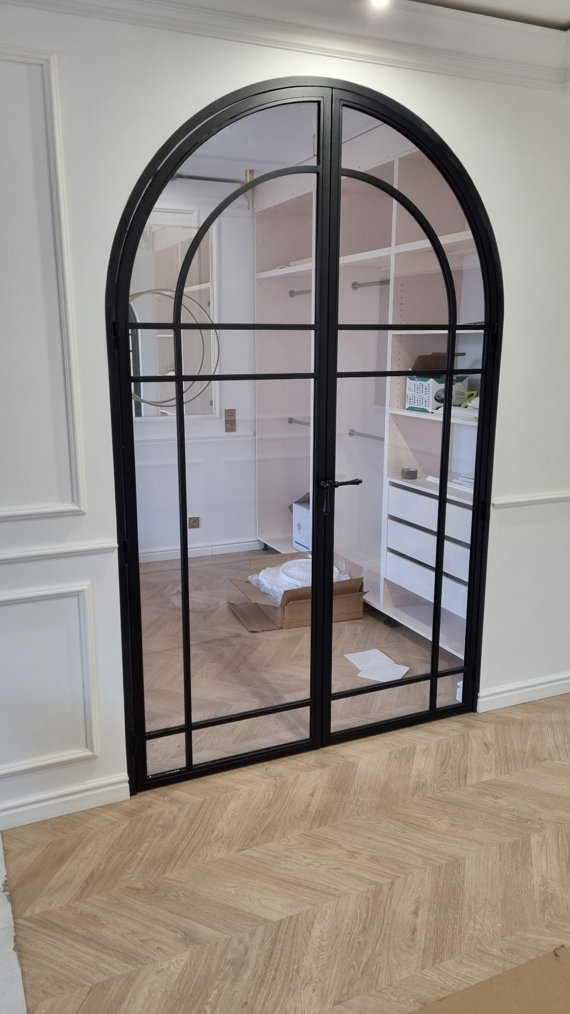 Dwuskrzydłowe drzwi łukowe z czarną, metalową ramą i szprosami, oddzielające garderobę od pokoju. Parkiet w jodełkę, białe ściany z dekoracyjną sztukaterią.