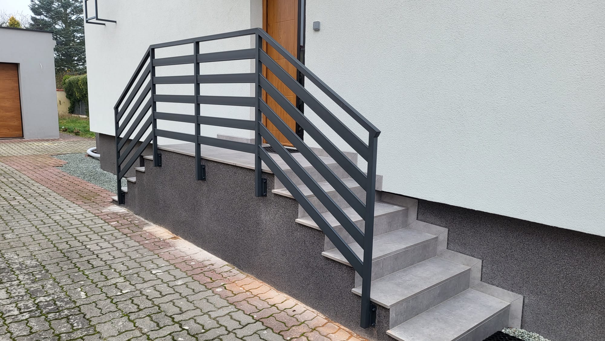 Nowoczesna, grafitowa balustrada schodowa z poziomymi szczeblami, zamontowana przy wejściu do domu z elewacją w odcieniach bieli i szarości. Widok z dołu, kostka brukowa na pierwszym planie.