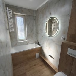 Mateusz Pencarski - Jasna łazienka z wanną, okrągłym lustrem z podświetleniem LED i płytkami imitującymi marmur oraz drewno. Widoczne baterie, grzejnik drabinkowy i toaleta.