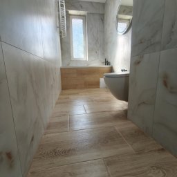 Mateusz Pencarski - Wykończona łazienka z beżowymi płytkami imitującymi drewno na podłodze i marmurkowymi na ścianach. Widoczna toaleta, grzejnik drabinkowy i okrągłe lustro.