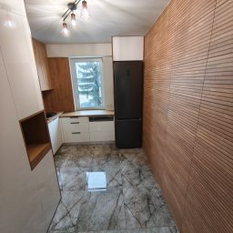 Mateusz Pencarski - Nowoczesna kuchnia z marmurową podłogą, drewnianą ścianą i czarną lodówką. Białe szafki kuchenne pod oknem. Minimalistyczne oświetlenie sufitowe.