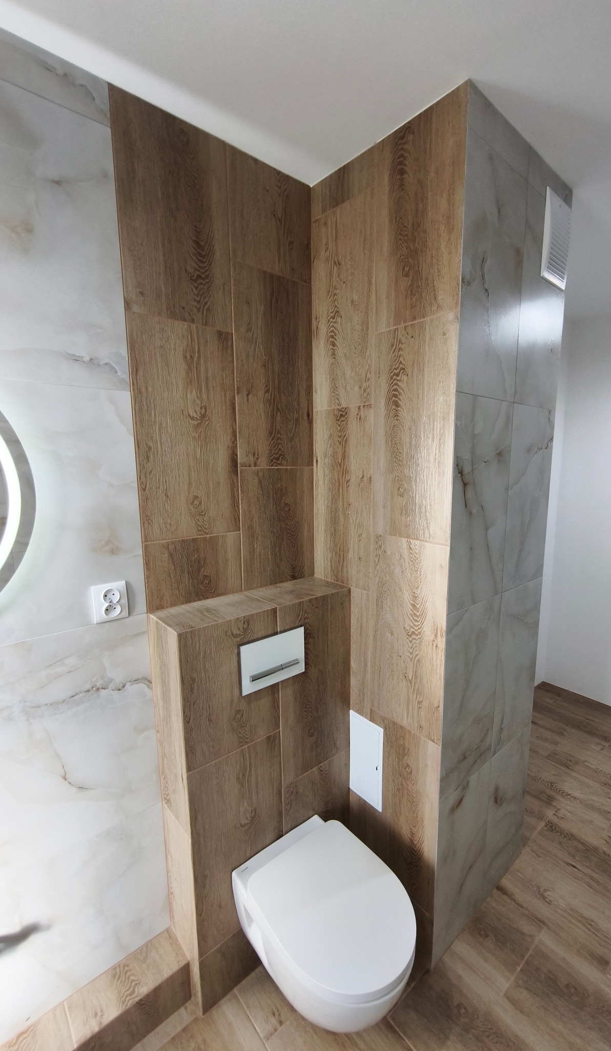 Nowoczesna toaleta podwieszana w wykończonej łazience. Brązowe płytki imitujące drewno na ścianach i podłodze, marmurowe płytki na ścianie z lustrem.