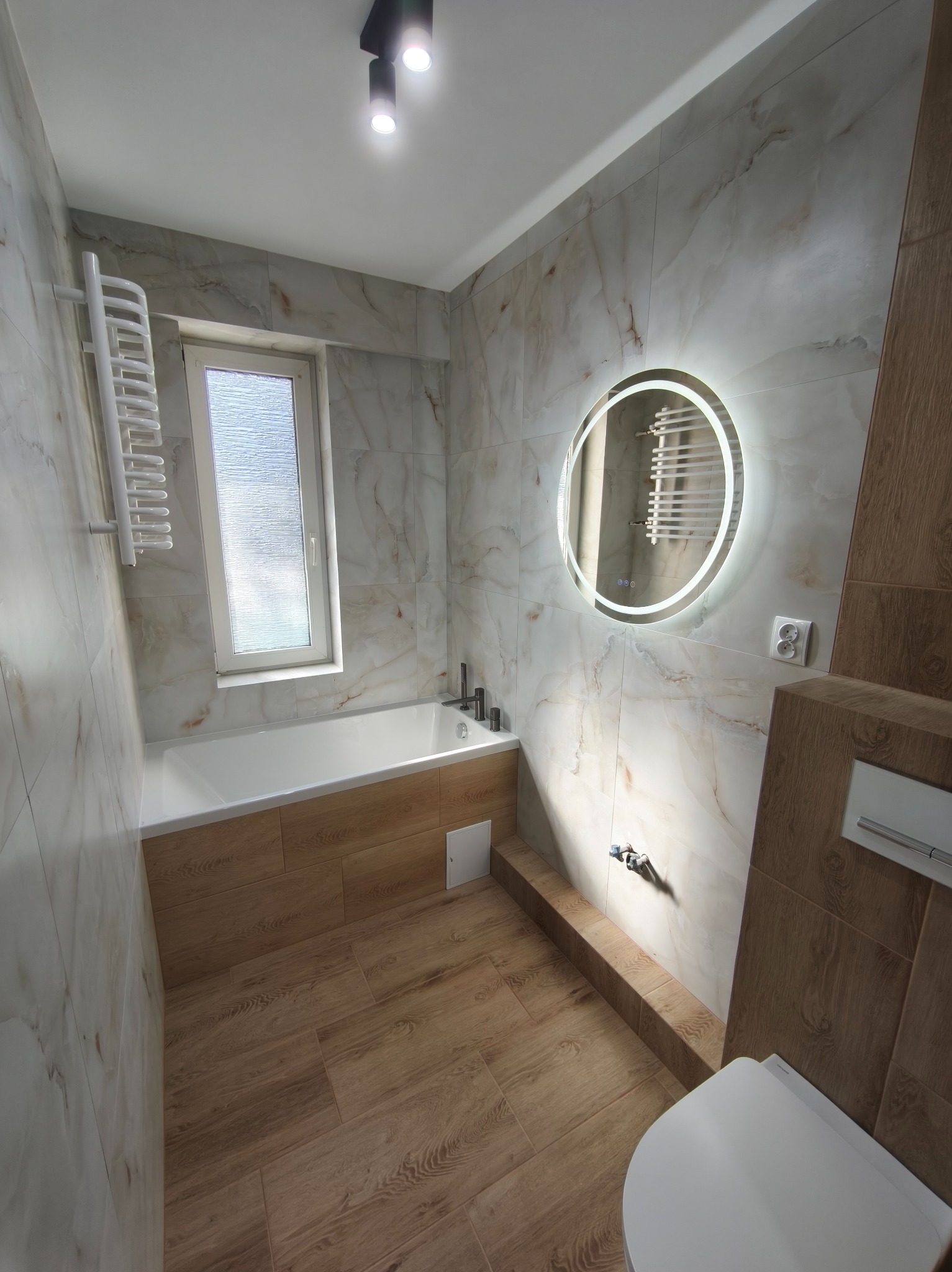 Jasna łazienka z wanną, okrągłym lustrem z podświetleniem LED i płytkami imitującymi marmur oraz drewno. Widoczne baterie, grzejnik drabinkowy i toaleta.