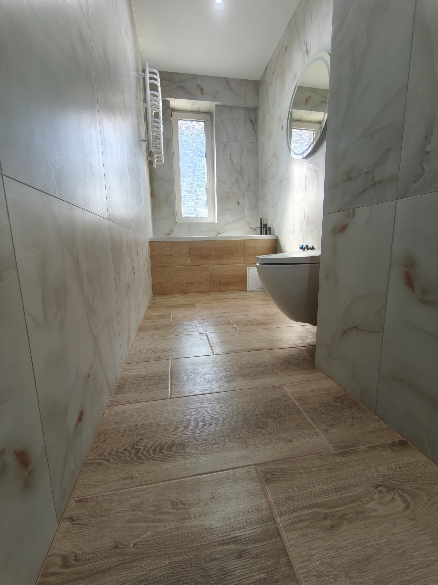Wykończona łazienka z beżowymi płytkami imitującymi drewno na podłodze i marmurkowymi na ścianach. Widoczna toaleta, grzejnik drabinkowy i okrągłe lustro.