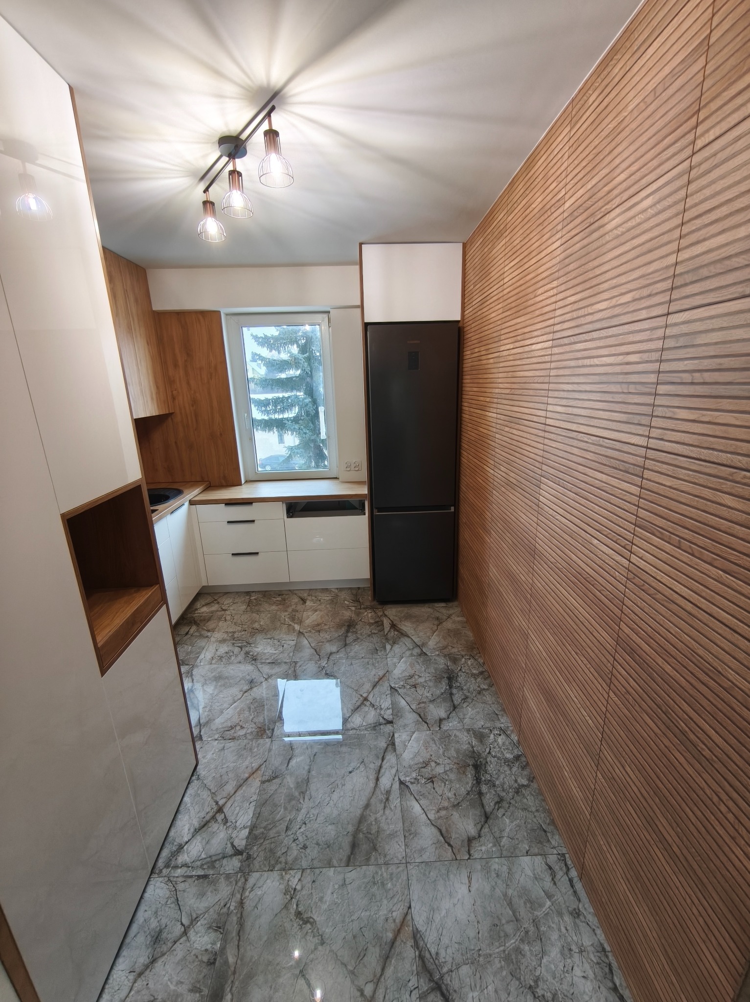 Nowoczesna kuchnia z marmurową podłogą, drewnianą ścianą i czarną lodówką. Białe szafki kuchenne pod oknem. Minimalistyczne oświetlenie sufitowe.