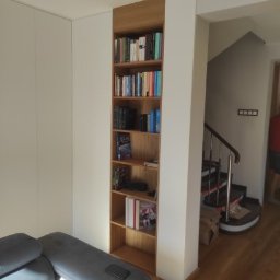 Pasja i oddanie - Wnęka z drewnianymi półkami na książki, wkomponowana w białą zabudowę, widoczna z perspektywy salonu z szarą sofą i fragmentem schodów.