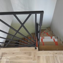 Pasja i oddanie - Nowoczesna balustrada schodowa z czarnego metalu o geometrycznym wzorze, widok z góry na drewniane schody zabezpieczone kartonem i taśmą. Jasne wnętrze.