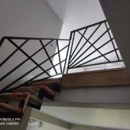 Pasja i oddanie - Nowoczesna balustrada schodowa z czarnymi, geometrycznymi elementami, widok z dołu, częściowo zakryte schody w trakcie montażu, jasne tło.