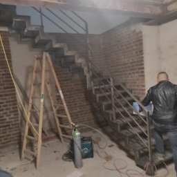 Pasja i oddanie - Montaż metalowych schodów z balustradą w surowym wnętrzu z ceglaną ścianą. Widoczna drabina i butla spawalnicza, sugerujące trwające prace wykończeniowe.
