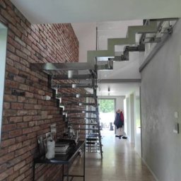 Pasja i oddanie - Nowoczesne schody metalowe z minimalistyczną konstrukcją, przylegające do ceglanej ściany w jasnym wnętrzu. Widoczne elementy dekoracyjne i oświetlenie.