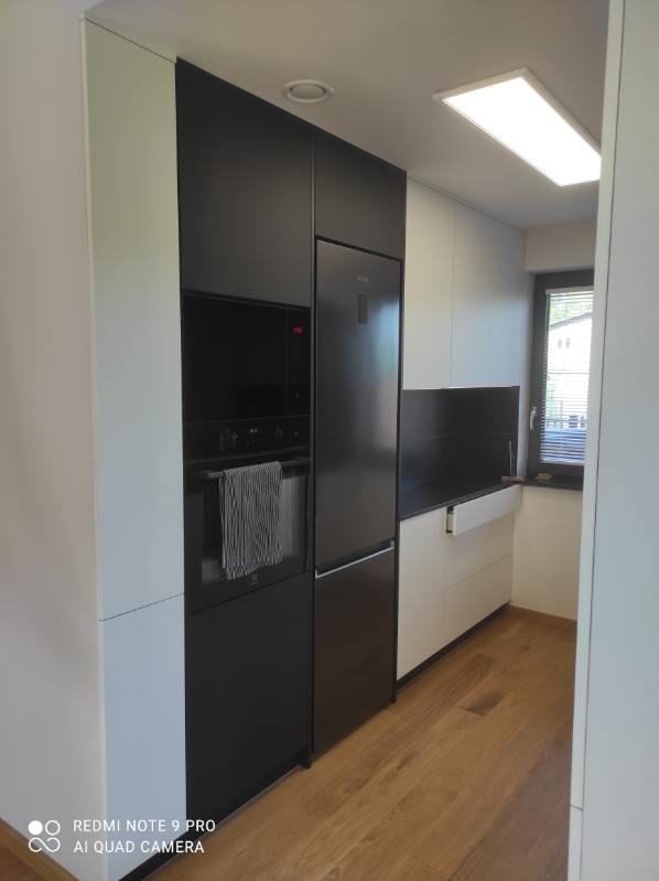 Nowoczesna kuchnia z zabudową w kolorach bieli i czerni. Minimalistyczny design, drewniana podłoga, sprzęt AGD w zabudowie. Oświetlenie sufitowe.