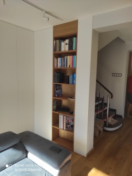 Wnęka z drewnianymi półkami na książki, wkomponowana w białą zabudowę, widoczna z perspektywy salonu z szarą sofą i fragmentem schodów.