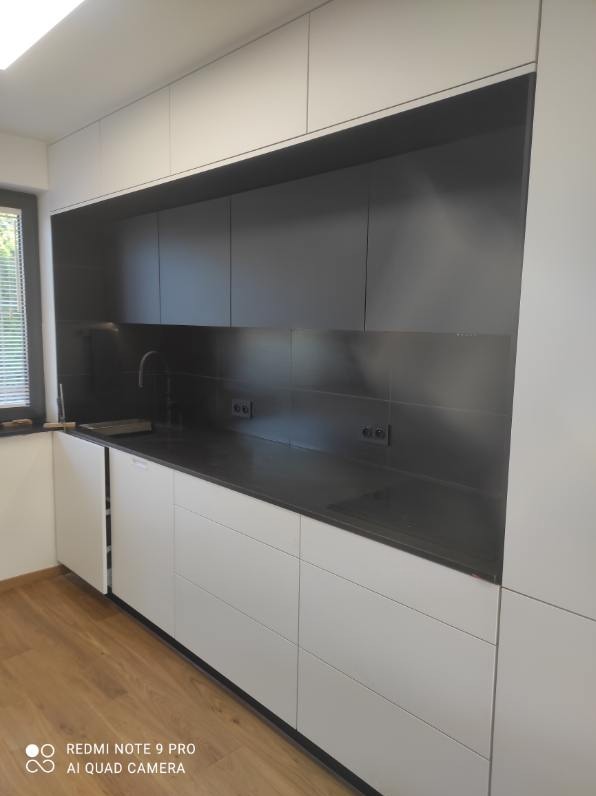 Nowoczesna kuchnia z białymi szafkami dolnymi i ciemnymi górnymi, czarny blat i ściana nad blatem, minimalistyczny design, widok na okno z żaluzjami.