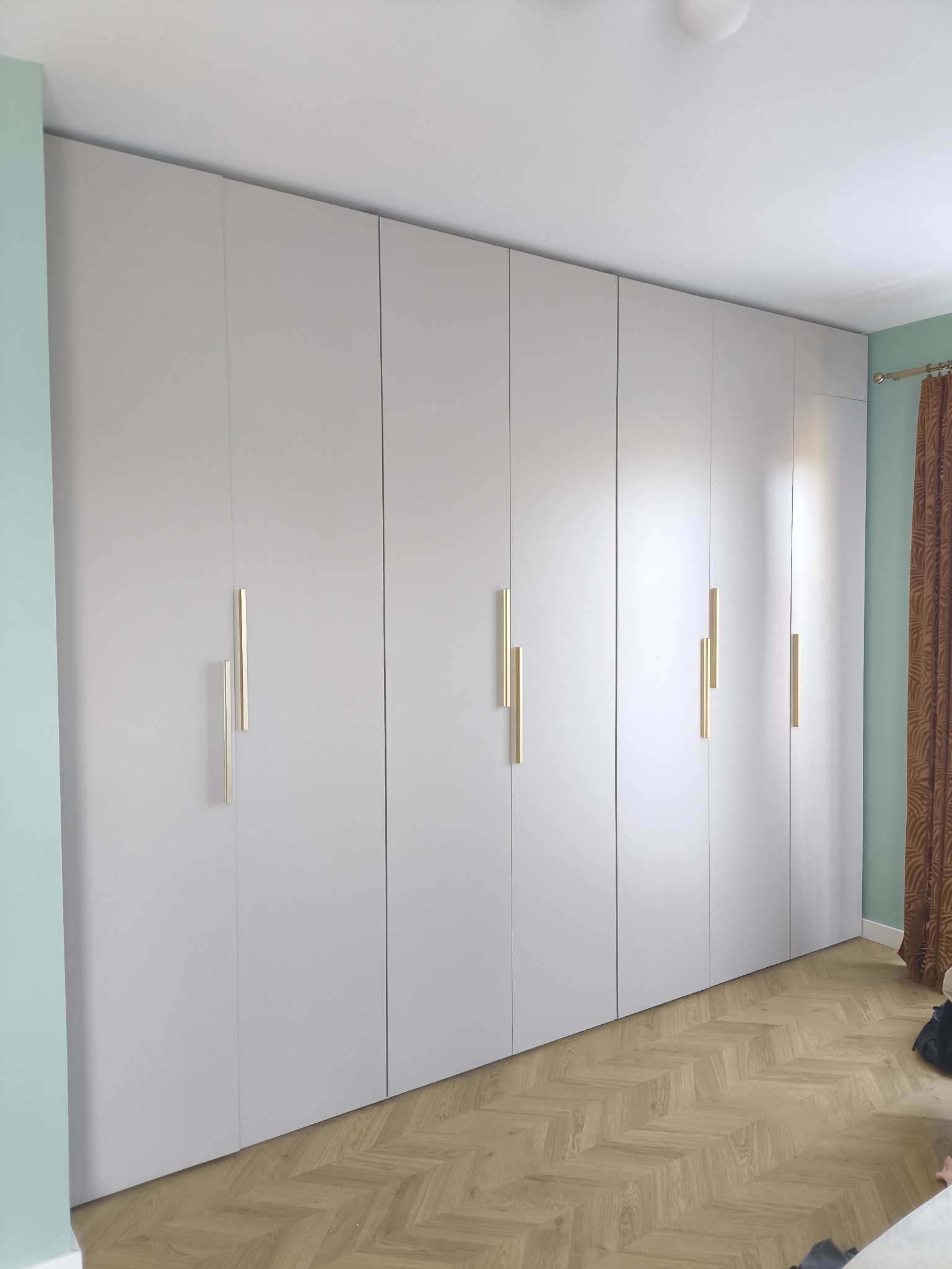 Jasnoszara szafa wnękowa z złotymi, pionowymi uchwytami na tle parkietu w jodełkę i miętowej ściany. Minimalistyczny design, nowoczesne wykończenie.