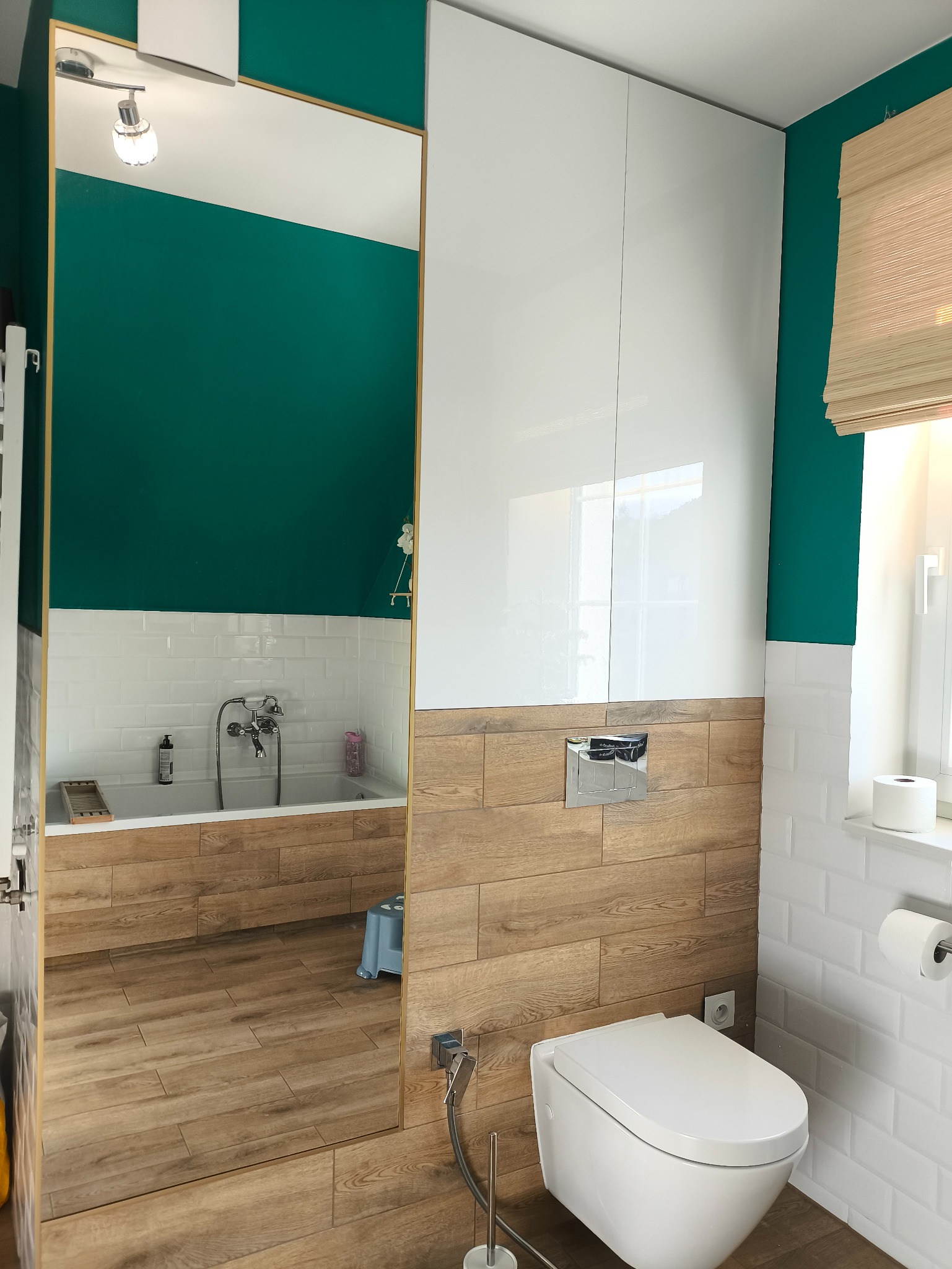 Nowoczesna łazienka z drewnopodobnymi płytkami i białymi szafkami. Lustro z widokiem na wannę i zieloną ścianę. Minimalistyczny design, jasne oświetlenie.