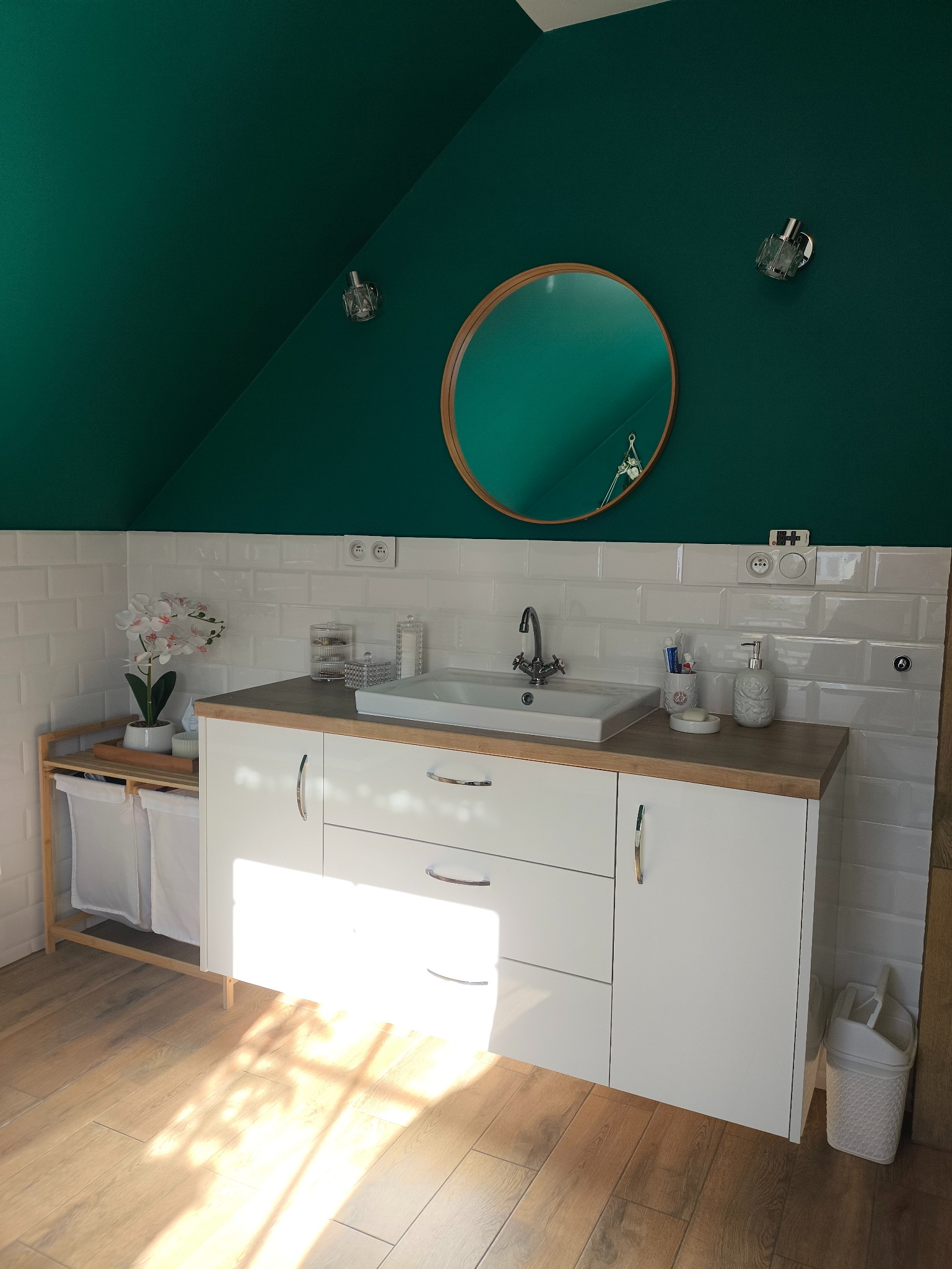 Nowoczesna łazienka na poddaszu z białą szafką pod umywalkę, drewnianym blatem i okrągłym lustrem. Ściany wyłożone białymi płytkami, a sufit pomalowany na intensywny zielony kolor.