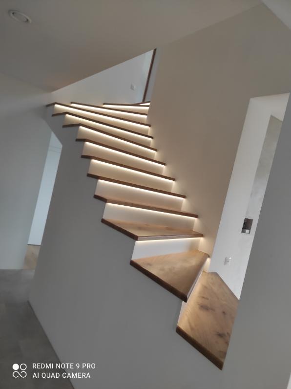 Nowoczesne schody z drewnianymi stopniami i subtelnym oświetleniem LED, wkomponowane w minimalistyczne, białe wnętrze. Widok z dołu, akcentujący geometrię i światło.