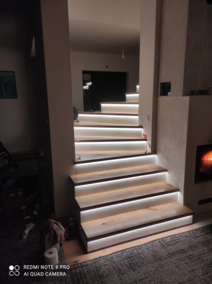 Nowoczesne schody z podświetleniem LED, drewniane stopnie, jasne wnętrze, widok z dołu, ciepłe światło, minimalistyczny design, element kominka w tle.