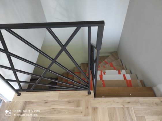 Nowoczesna balustrada schodowa z czarnego metalu o geometrycznym wzorze, widok z góry na drewniane schody zabezpieczone kartonem i taśmą. Jasne wnętrze.