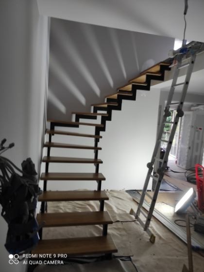 Nowoczesne schody z drewnianymi stopniami i czarną konstrukcją, widoczne podczas montażu, z drabiną opartą o ścianę. Minimalistyczny design, surowe wykończenie wnętrza.