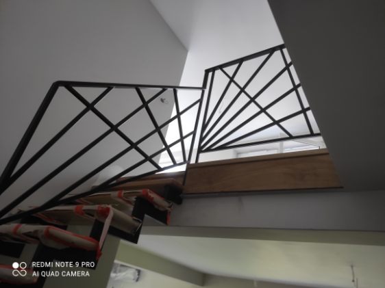 Nowoczesna balustrada schodowa z czarnymi, geometrycznymi elementami, widok z dołu, częściowo zakryte schody w trakcie montażu, jasne tło.